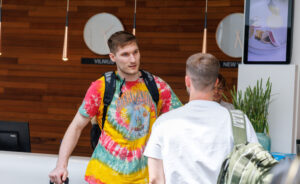 Traveler with a colorful tie-dye shirt and backpack talks to a staff member at a white reception desk in a modern lobby.