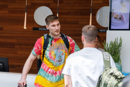 Traveler with a colorful tie-dye shirt and backpack talks to a staff member at a white reception desk in a modern lobby.