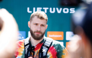 Bearded man in a tie-dye shirt wearing a harness, being interviewed with sponsor logos blurred in the background.