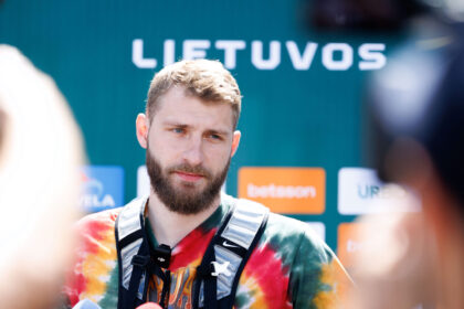 Bearded man in a tie-dye shirt wearing a harness, being interviewed with sponsor logos blurred in the background.