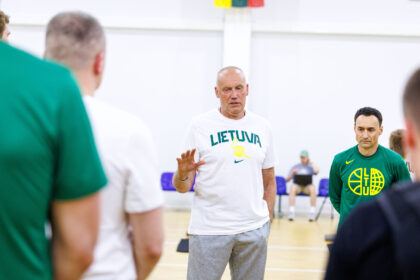 Central man in a white Lietuva T-shirt addressing a group in a gym as others in green shirts listen nearby
