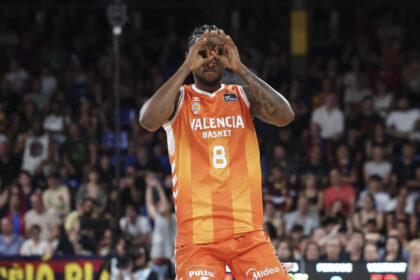 Basketball player in orange Valencia Basket uniform on court making binocular gesture with hands over eyes, crowd blurred behind.