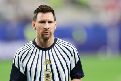 Soccer player in a white and navy striped jersey with AFA crest, standing on a green field with a blurred stadium background