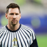 Soccer player in a white and navy striped jersey with AFA crest, standing on a green field with a blurred stadium background