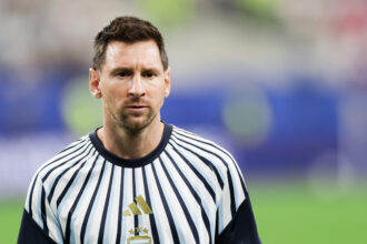 Soccer player in a white and navy striped jersey with AFA crest, standing on a green field with a blurred stadium background
