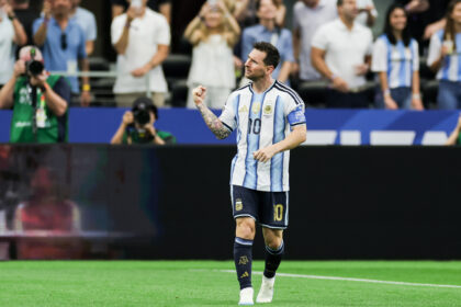 Argentine soccer player in a light-blue striped jersey celebrating on the field with a clenched fist raised, fans in the stands behind him