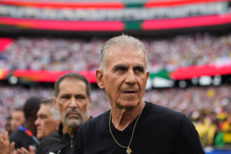 Close-up of a man in a black shirt wearing a gold pendant, with a blurred stadium crowd in the background.