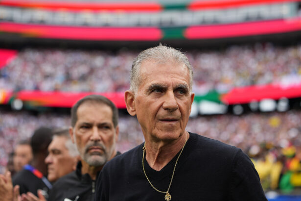Close-up of a man in a black shirt wearing a gold pendant, with a blurred stadium crowd in the background.