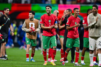 Moroccan national team players in red jerseys and green shorts stand on a soccer pitch, smiling and clapping after a match.