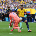 Soccer match action as a yellow-jersey attacker (#11) challenges a pink-clad goalkeeper for the ball, with defenders nearby and a crowded stadium behind.