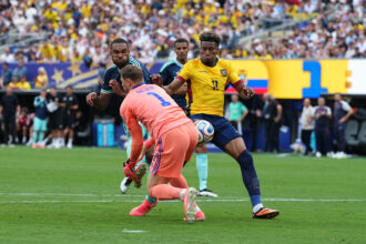 Soccer match action as a yellow-jersey attacker (#11) challenges a pink-clad goalkeeper for the ball, with defenders nearby and a crowded stadium behind.