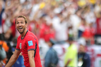 Close-up of a male soccer player in a red jersey celebrating on the field with a cheering crowd in the stands behind him.