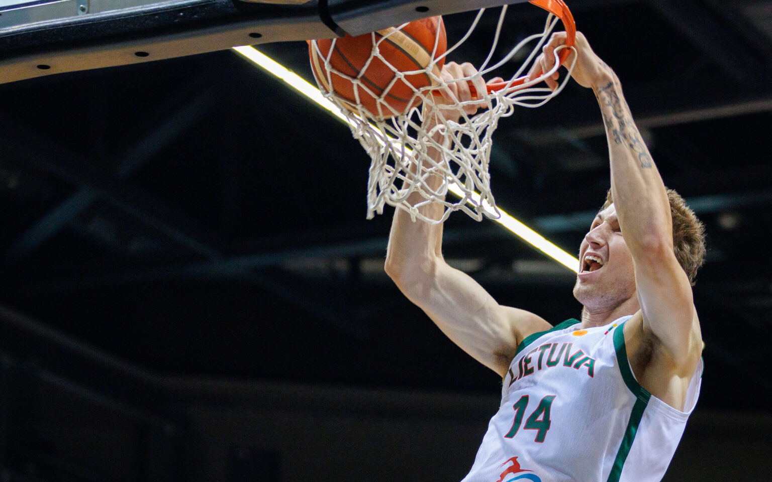 Basketball player in white-and-green jersey #14 dunks the ball through the hoop, gripping the rim with both hands as the net hangs down.