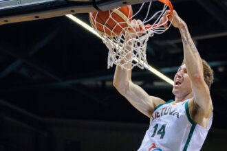 Basketball player in white-and-green jersey #14 dunks the ball through the hoop, gripping the rim with both hands as the net hangs down.