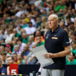 Basketball coach standing on the sideline, clipboard in hand, watching a game with a crowded arena in the background.