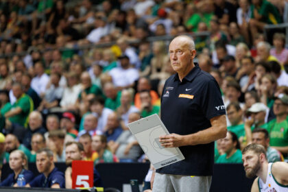 Basketball coach standing on the sideline, clipboard in hand, watching a game with a crowded arena in the background.
