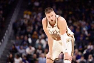 Basketball player in a white-brown uniform crouches on the court during a game, blurred crowd in the background.