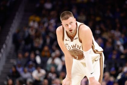 Basketball player in a white-brown uniform crouches on the court during a game, blurred crowd in the background.