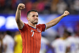 Soccer player in a red striped jersey raising both fists in celebration on the field, crowd blurred in the background, mid-shot.