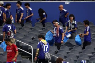 Group of soccer fans in blue jerseys walking through empty stadium seating, carrying blue bags and wearing face paint and scarves.