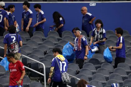 Group of soccer fans in blue jerseys walking through empty stadium seating, carrying blue bags and wearing face paint and scarves.