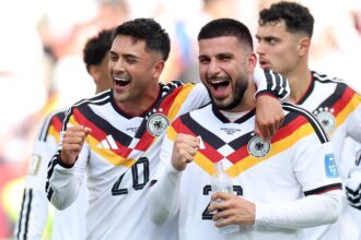 German footballers celebrate together, arms around each other, smiling in German national team jerseys with black-red-yellow diagonal stripes.