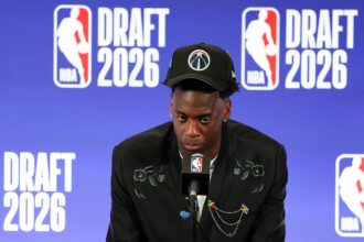 Young man in a black suit and cap at a press conference, speaking into a microphone with a blue NBA Draft backdrop that reads 'DRAFT 2020' behind him.