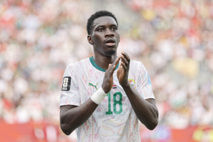 Soccer player in a light patterned jersey applauding on the field, crowd blurred in the stands behind him, World Cup 2026 badge visible on sleeve.