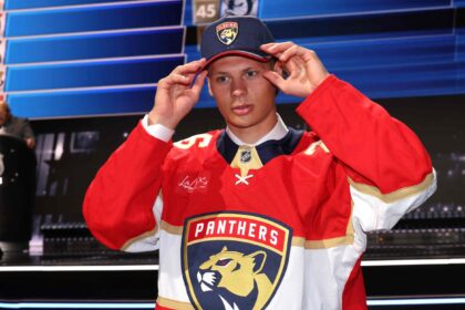 Hockey player in a red Florida Panthers jersey adjusts a cap during a public event on a blue-lit stage.