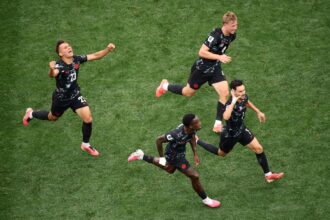 Aerial view of soccer players in black uniforms sprinting across a green grass field, celebrating a goal.