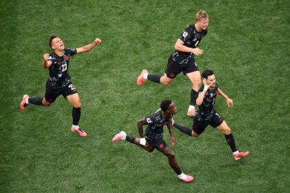 Aerial view of soccer players in black uniforms sprinting across a green grass field, celebrating a goal.