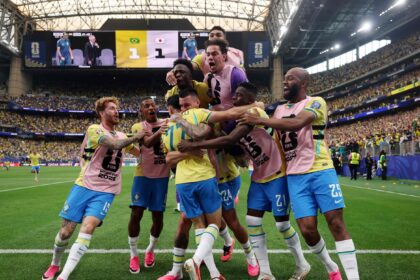 Brazilian soccer players in yellow jerseys celebrate a goal on the field with pink training bibs and a crowded stadium in the background.