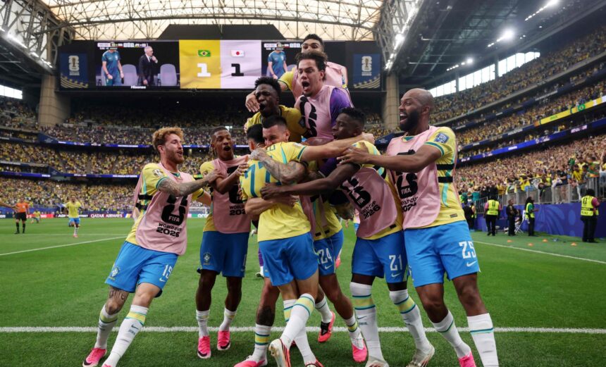 Brazilian soccer players in yellow jerseys celebrate a goal on the field with pink training bibs and a crowded stadium in the background.