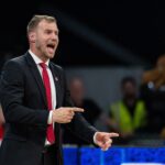 Coach in a black suit and red tie shouting instructions on the court with a crowd behind him.