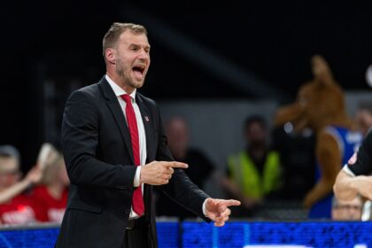 Coach in a black suit and red tie shouting instructions on the court with a crowd behind him.