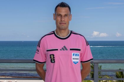 Male soccer referee in a pink FIFA jersey standing on a seaside boardwalk with the ocean behind him.