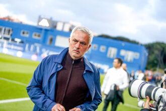 Mid-shot of a mature man in a blue jacket on a football field, looking toward the camera.