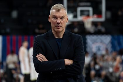 Man with short gray hair in a dark blazer stands with arms crossed in a gymnasium, basketball hoop blurred in the background.