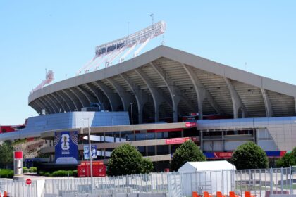 Exterior view of a large stadium with curved concrete arches and white construction scaffolding on the roof, FIFA World Cup 2026 banners visible in the foreground.