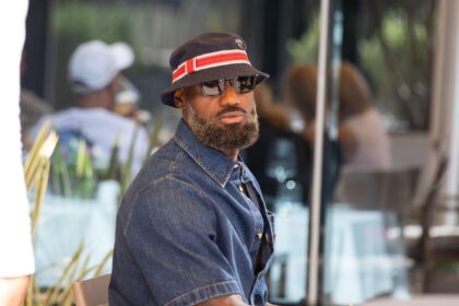 Man wearing a denim shirt and a dark bucket hat with a red stripe, sunglasses, and a gray beard sits at an outdoor cafe.