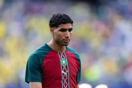 Portrait of a man with short, curly black hair wearing a red and teal Moroccan national team jersey, looking down, with a blurred stadium crowd in the background.