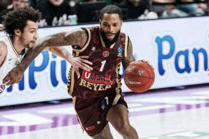 Basketball player in maroon uniform dribbles the ball while defended by an opponent on a purple court with advertising boards in the background.