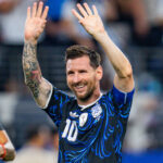 Soccer player in blue and black jersey raises both hands in celebration, smiling with tattoos visible on his left arm.