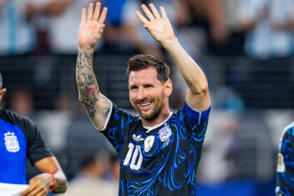Soccer player in blue and black jersey raises both hands in celebration, smiling with tattoos visible on his left arm.