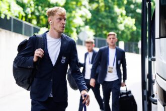 Young man in a navy blazer with a backpack walks toward a bus, with fellow suited students in the background.