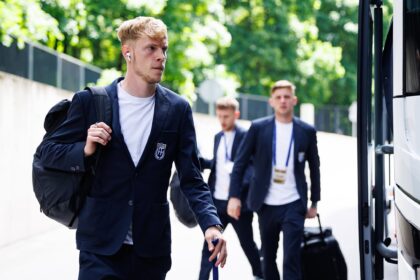 Young man in a navy blazer with a backpack walks toward a bus, with fellow suited students in the background.