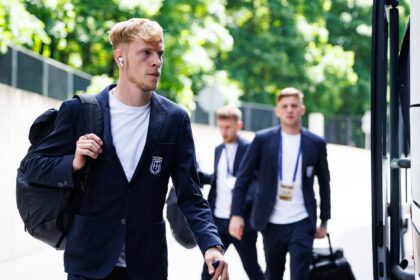 Young man in a navy blazer with a backpack walks toward a bus, with fellow suited students in the background.