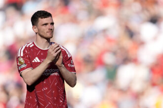 Soccer player in a red jersey clapping on the field with a blurred stadium crowd in the background.