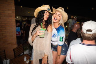 Two women with straw hats posing and smiling at a nighttime party, one holding a green cup with a straw.