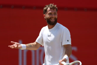 A man in a white shirt smiles while extending his left hand, holding a tennis racket in his right hand against a red background stadium wall.